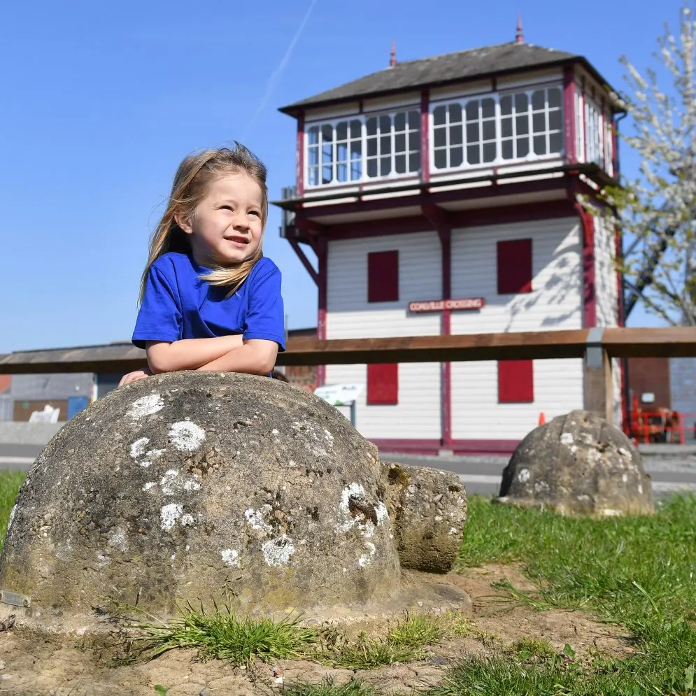 Young girl in blue shirt posing on lichen-covered stone with heritage colliery tower behind her.