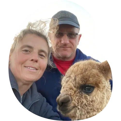 Two people smiling at the camera with a brown alpaca in the foreground.