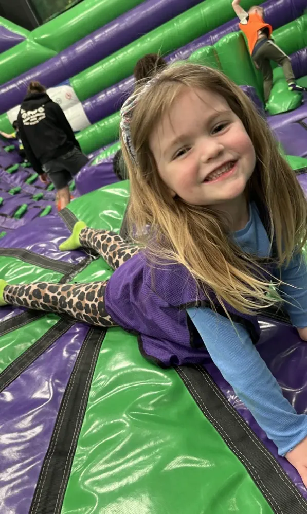 Young child smiling on a green and purple inflatable bouncy castle with other children playing behind.