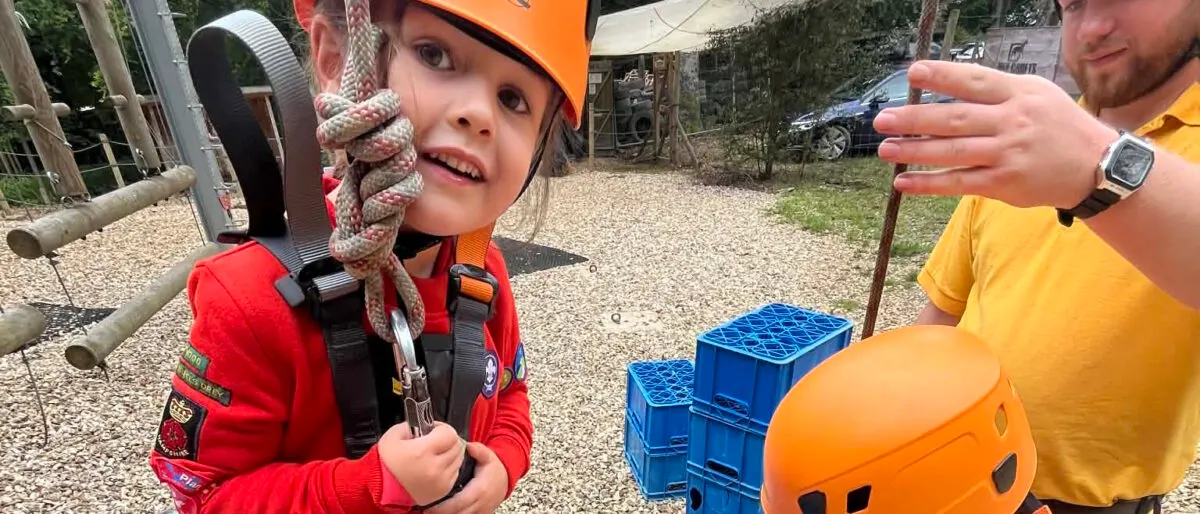 Young child in safety harness and orange helmet being instructed by an adult during an outdoor climbing or adventure activity.