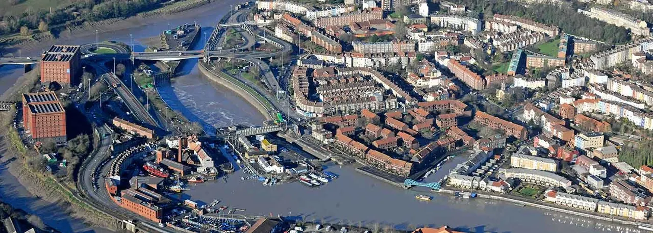 Aerial view of a riverside industrial area with historic brick buildings, waterfront developments, ships, and a curved river flowing through an urban landscape.