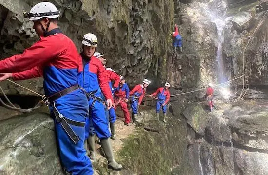 Group of people in safety gear canyoning through a narrow rocky gorge with a waterfall visible ahead.