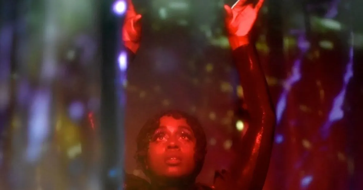 Person with arms raised, bathed in red stage lighting at a nighttime event or performance.