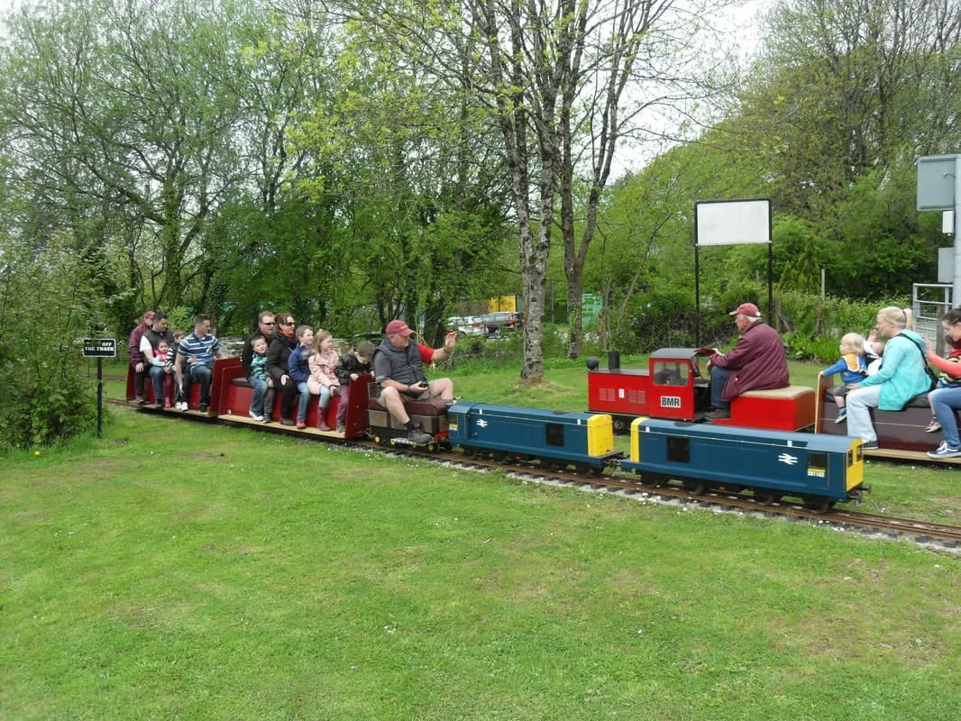 Children and adults riding a miniature railway train through a green park surrounded by trees.