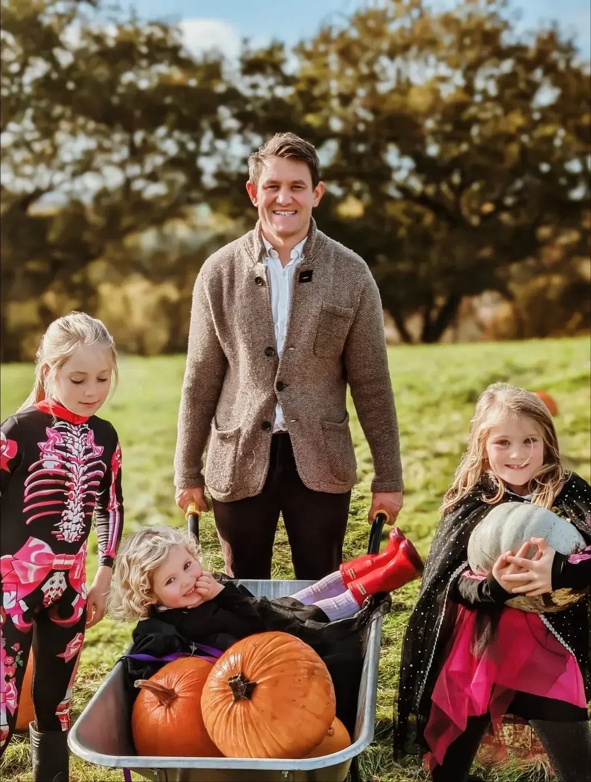 An adult and three young children in Halloween costumes stand in a field with a wheelbarrow full of pumpkins, trees visible in the background.
