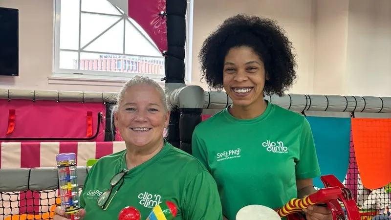 Two staff members in green uniforms smile at an indoor sports facility with colourful climbing holds and equipment visible behind them.