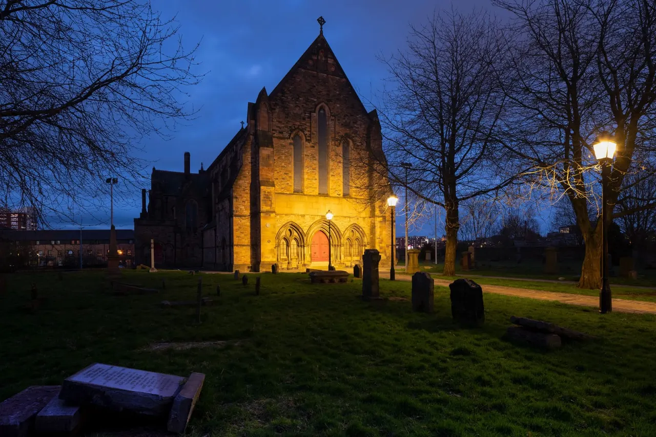 Historic stone church illuminated at dusk, surrounded by gravestones and bare trees in a churchyard setting.