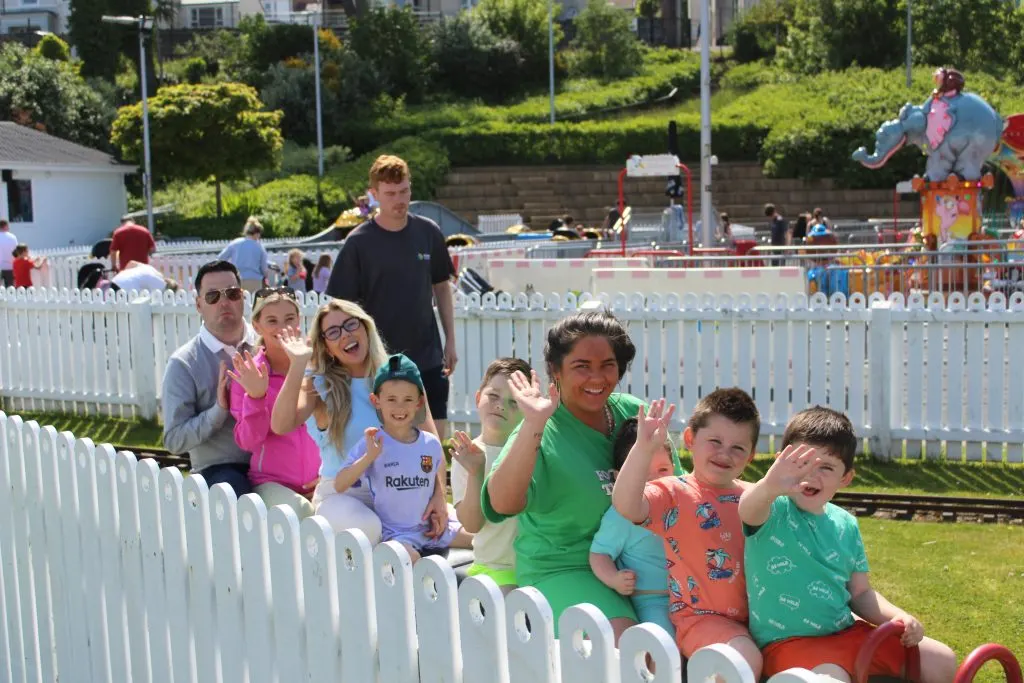 Adults and children waving at the camera while sitting by a white fence at an outdoor amusement park on a sunny day.