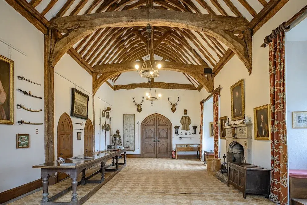 Historic timber-framed great hall with exposed wooden beams, period furniture, mounted deer heads, and decorated walls displaying weapons and portraits.