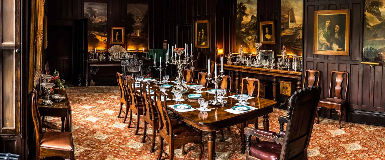 Ornate dining room with long wooden table set for a formal meal, surrounded by period furniture and adorned with paintings and candlelit chandeliers.