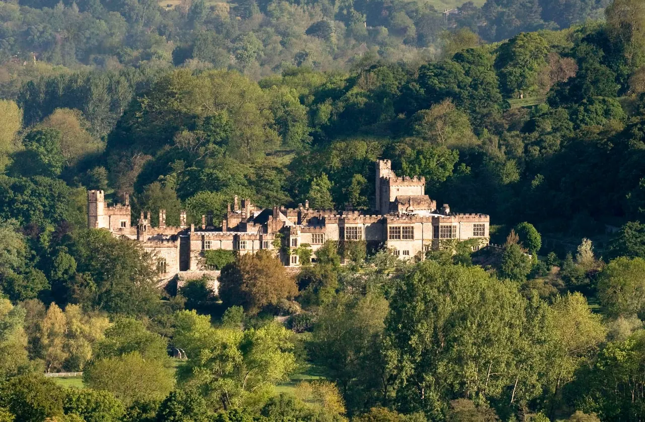 Historic stone castle and mansion nestled among dense green woodland and trees in a rural valley.