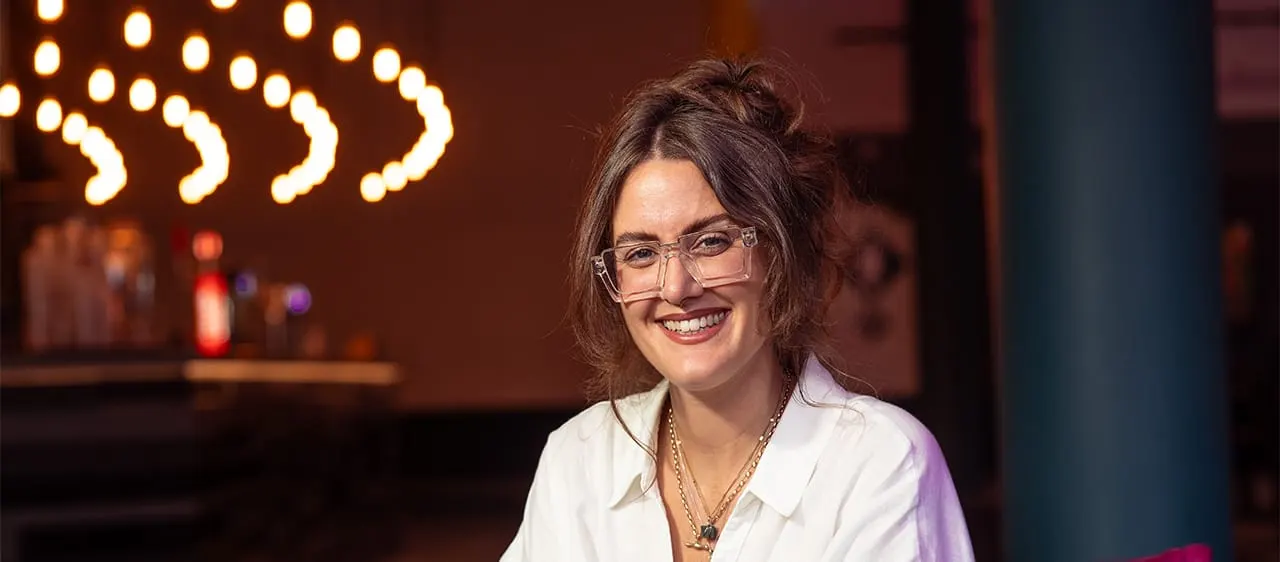 Woman wearing glasses and white shirt smiling at bar with warm neon lighting in background.
