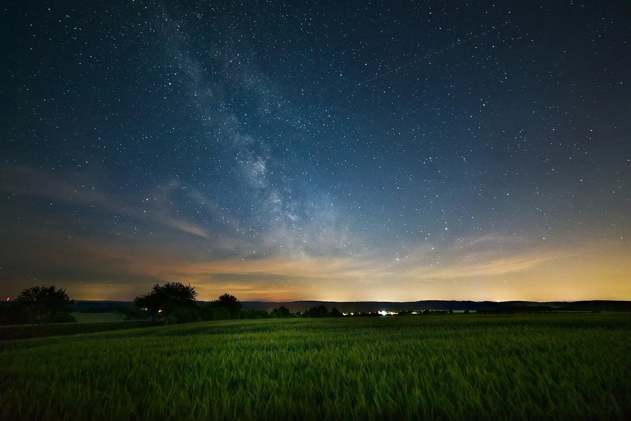 Milky Way and stars visible over a rural landscape with fields and distant trees at dusk.
