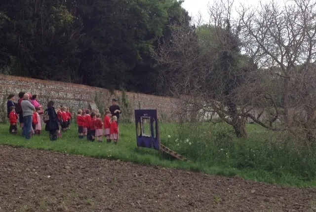 Group of children in red uniforms gathered on a grassy field beside a brick wall and bare trees.