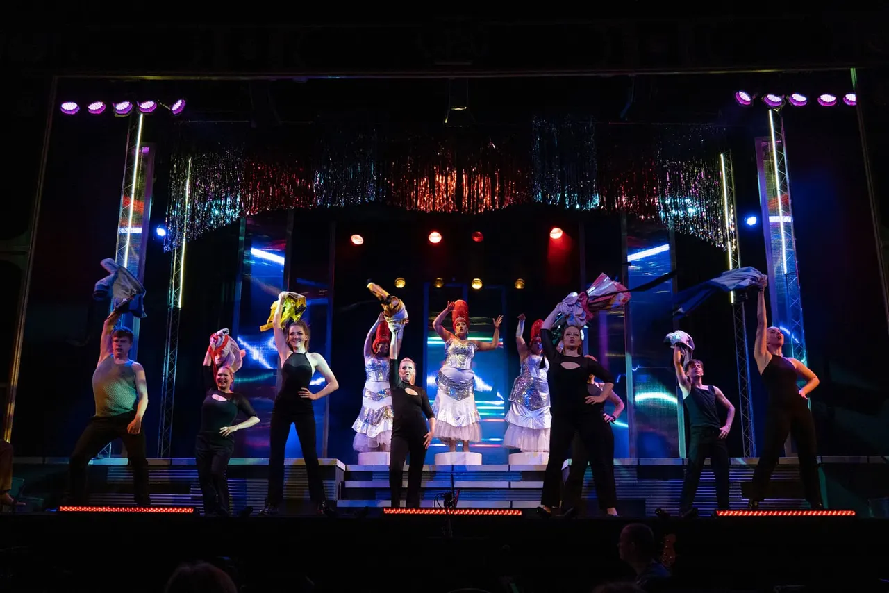 Performers in sequined costumes dance on an illuminated stage with neon lighting and hanging decorative elements during an evening show.