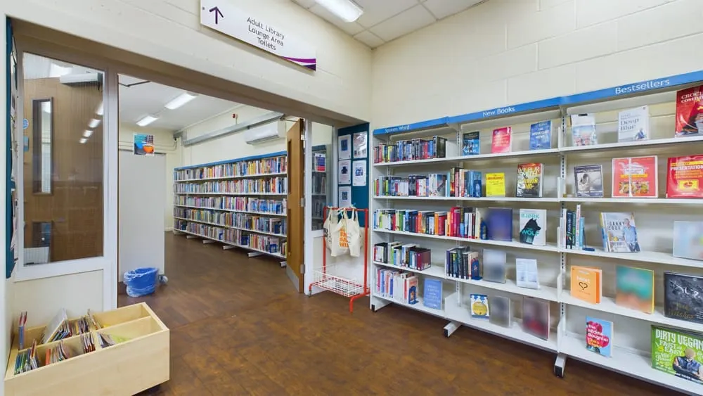 Interior of a library with shelving units displaying books and publications, a checkout desk area, and directional signage.