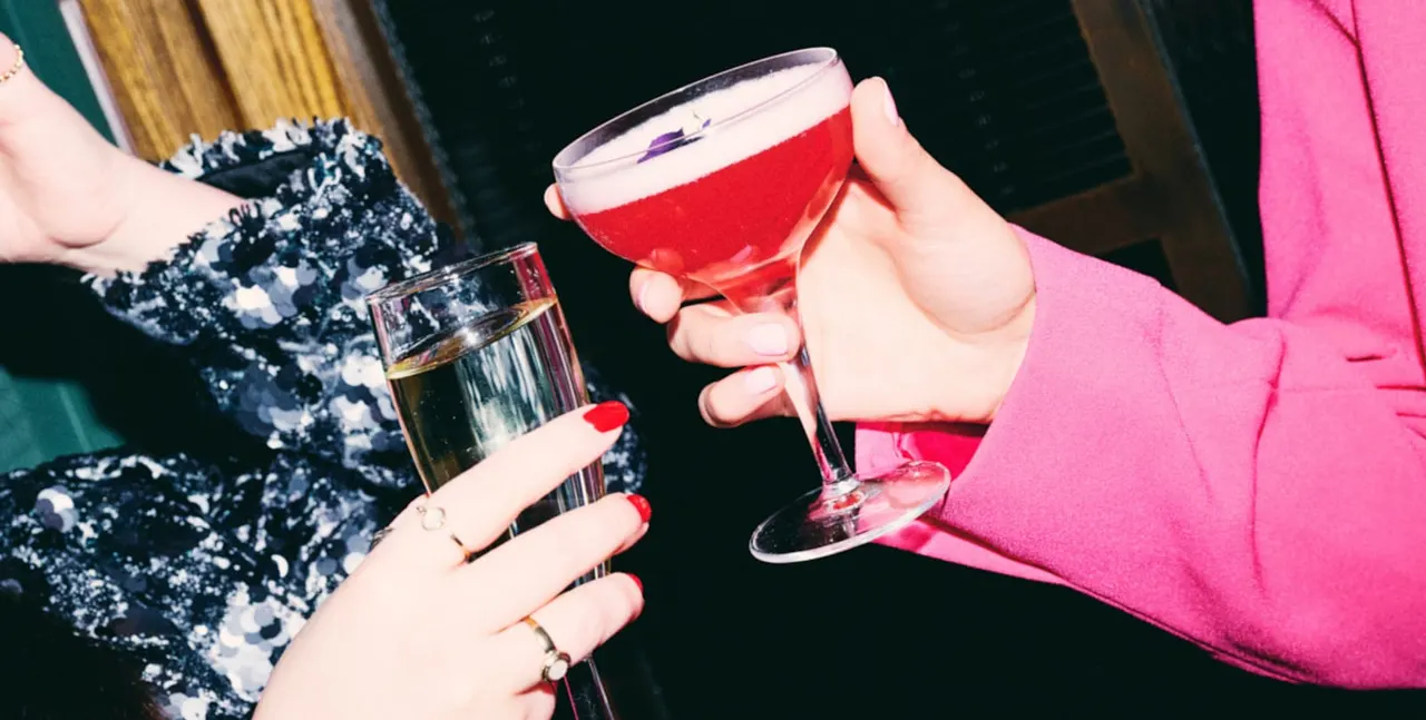 Two people in colourful outfits holding cocktails and a glass of sparkling wine at a social gathering.