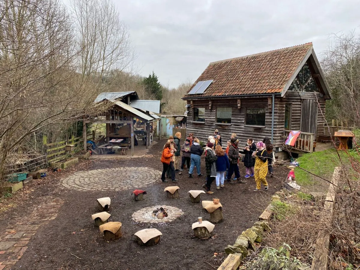 Group of children and adults gathered around a campfire with rustic wooden buildings and bare trees at an outdoor education space.