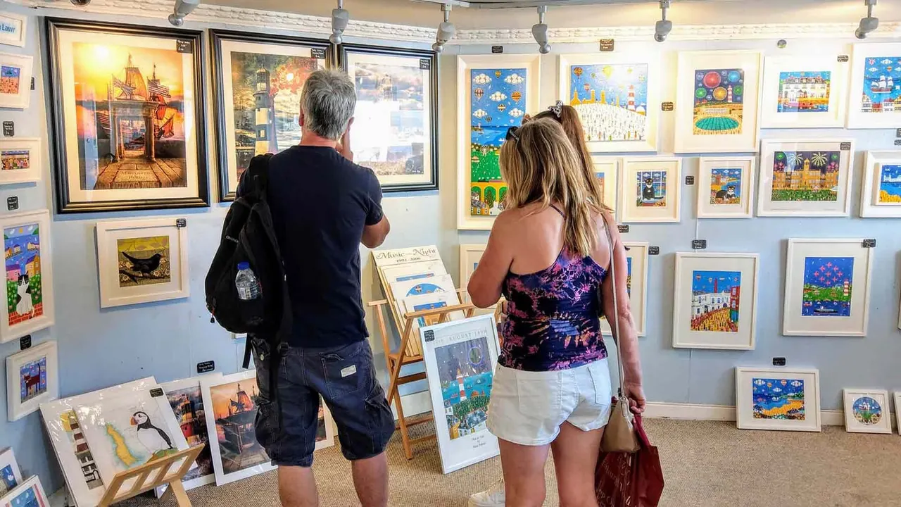Two visitors viewing colourful framed artworks and prints displayed on gallery walls and easels.