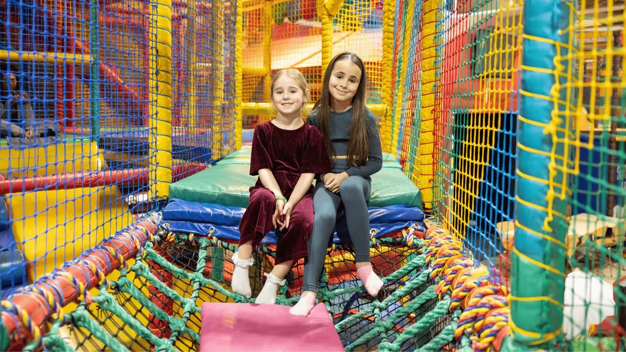 Two young children sitting together in a colourful indoor play structure with netting and foam elements.