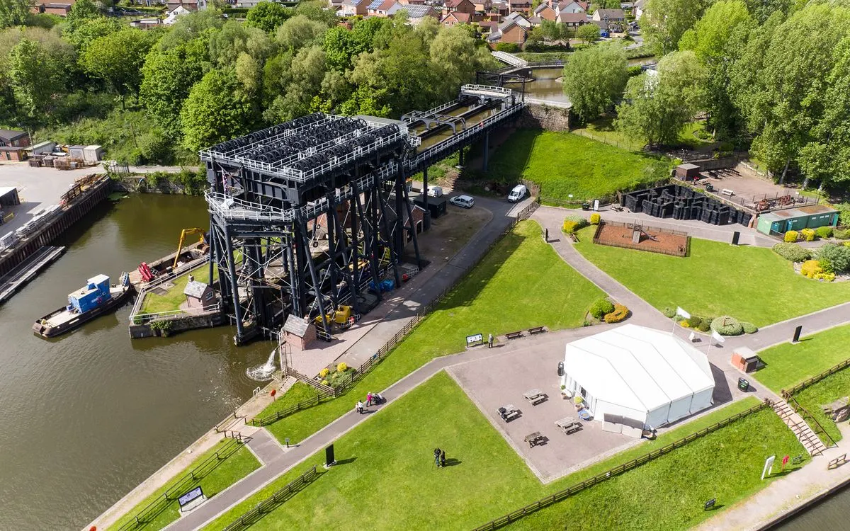 Aerial view of a historic boat lift structure spanning a canal with surrounding green lawns, visitor facilities, and residential buildings beyond.
