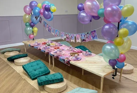 A children's party room decorated with colourful balloons and bunting, featuring a long table set with pink tableware and cushioned seating areas on the floor.