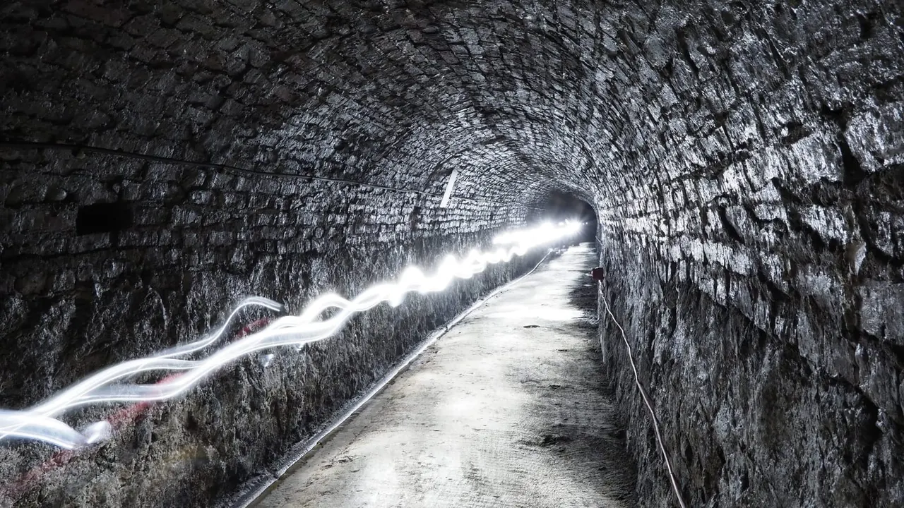 Stone tunnel with footpath and illuminated water channel running alongside, light trails visible on water surface.