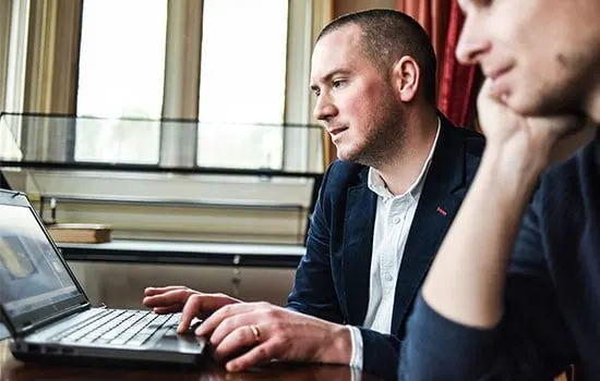 Two people work together at a laptop in an indoor setting with large windows.