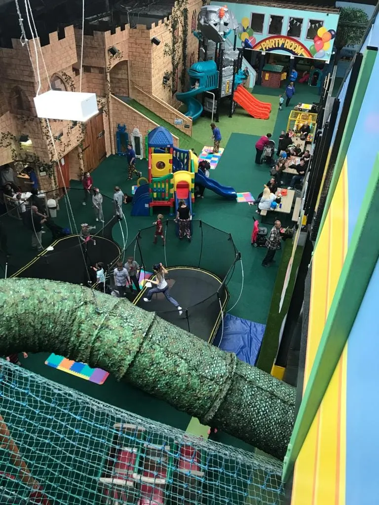 Overhead view of an indoor play centre with children playing on climbing structures, slides, trampolines and colourful equipment across a green floor.