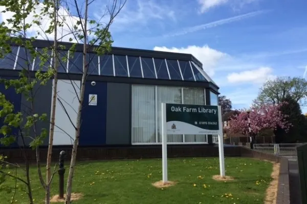 Modern black building with "Oak Farm Library" signage set in a green park with young trees and flowering shrubs under blue sky.