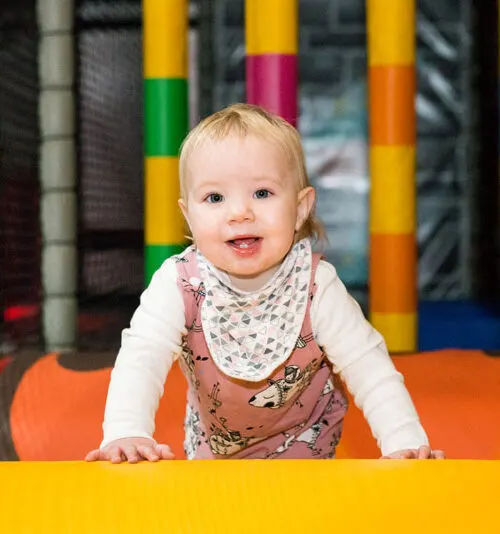 A toddler in a pink dress and white shirt smiles while crawling on colourful play equipment indoors.