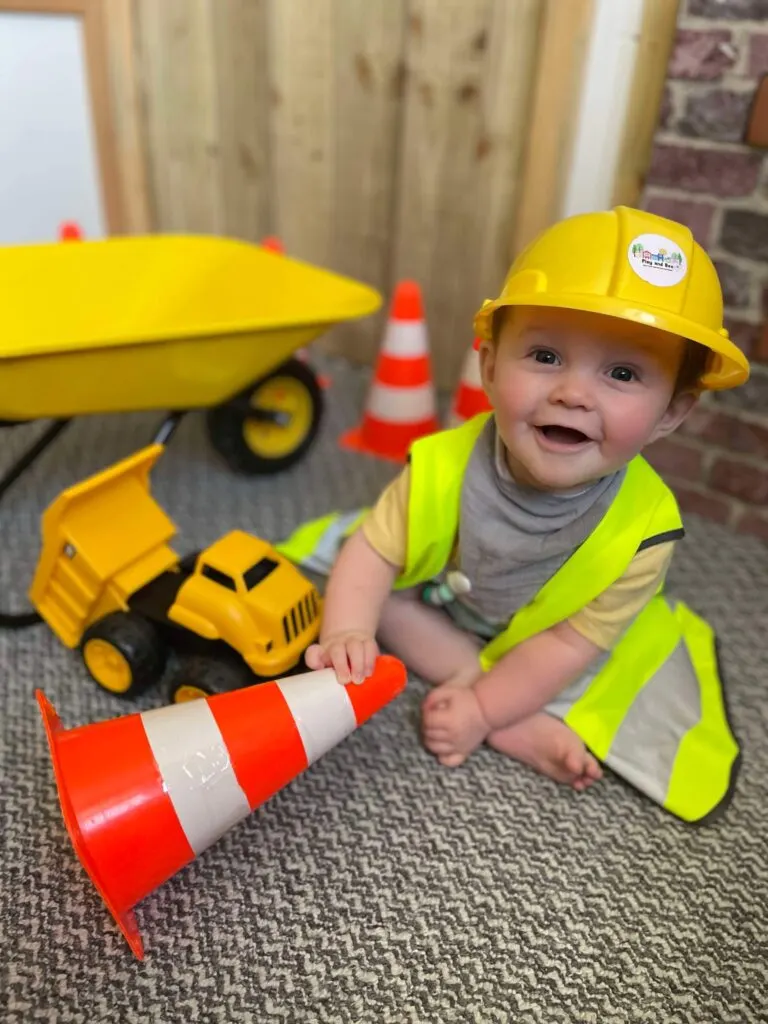 Young child wearing construction gear and hard hat playing with toy vehicles and traffic cones.