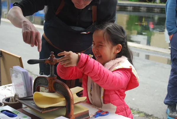 A child in a pink jacket operates a printing press with guidance from an adult at an outdoor demonstration.
