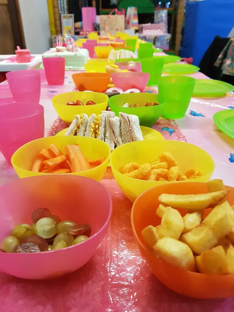 Children's party table set with colourful bowls of food including sandwiches, crisps, fruit and vegetables.