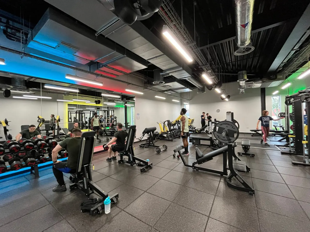 Gym interior with multiple members using various exercise equipment including weights, exercise bikes, and machines under colourful ceiling lighting.