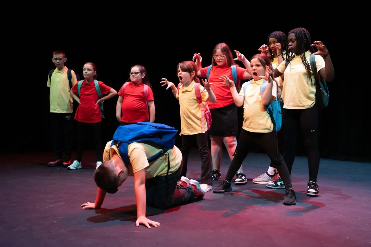 Child performing a backflip on stage whilst other children in coloured t-shirts watch and react with enthusiasm.