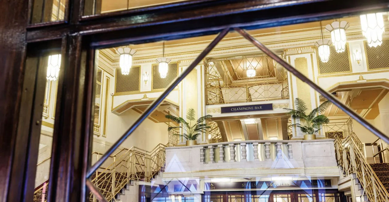 Art deco interior with grand staircase, ornate gold railings, pendant lights and a champagne bar sign visible on the mezzanine level.