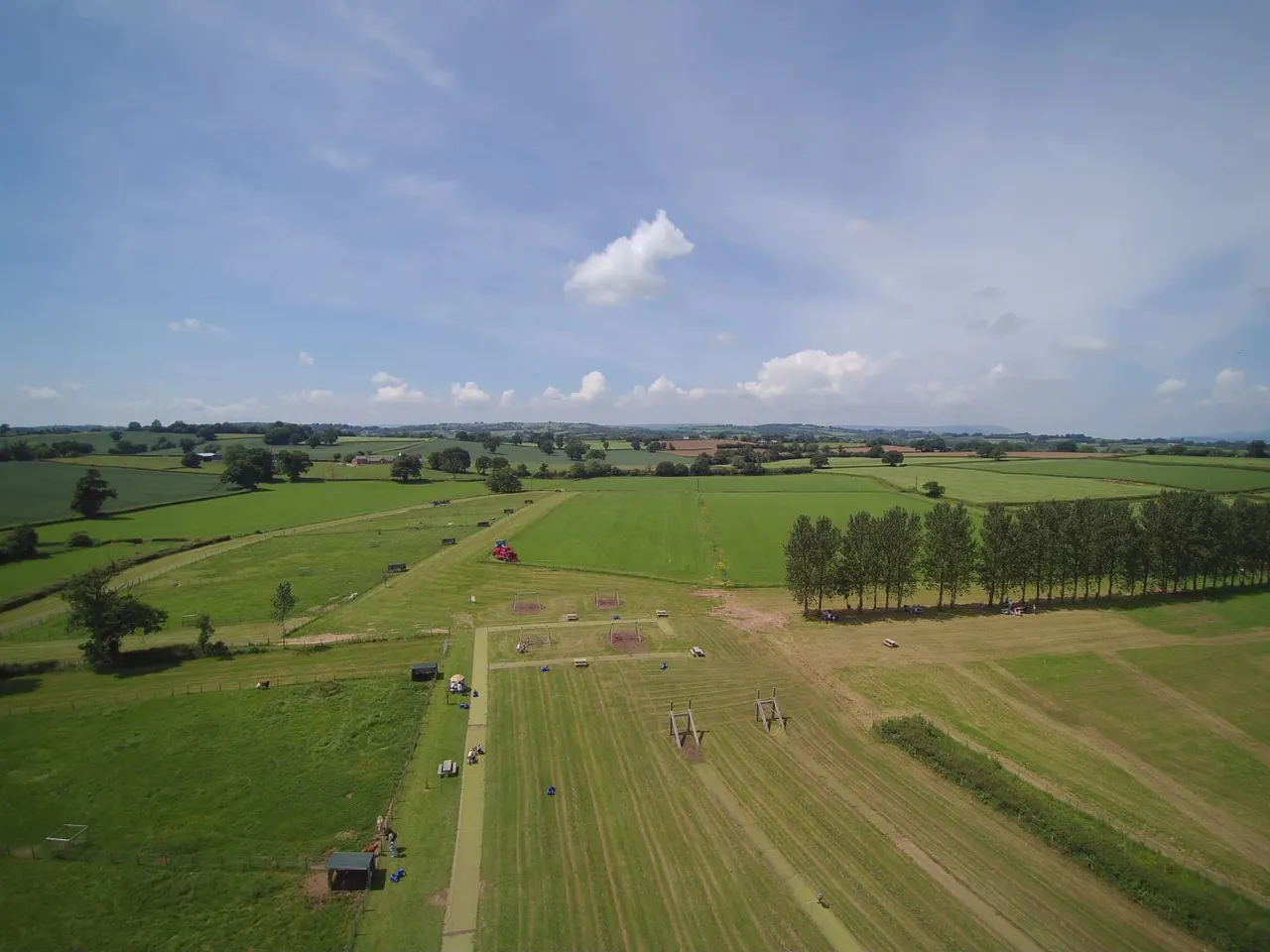 Aerial view of a farm with green fields, jump obstacles, and spectators gathered along a central pathway under a blue sky.