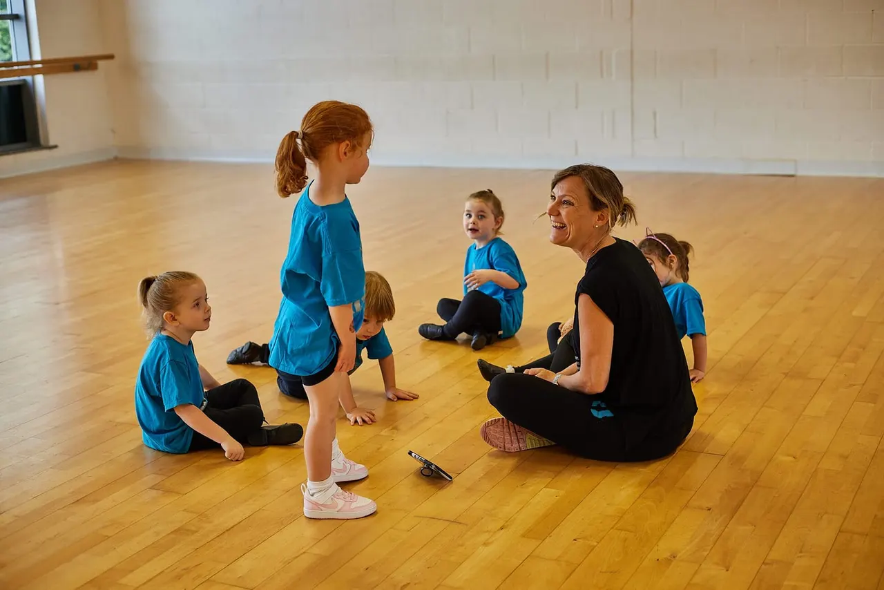 An instructor leads a group of young children in a dance or movement class in a wooden-floored studio.
