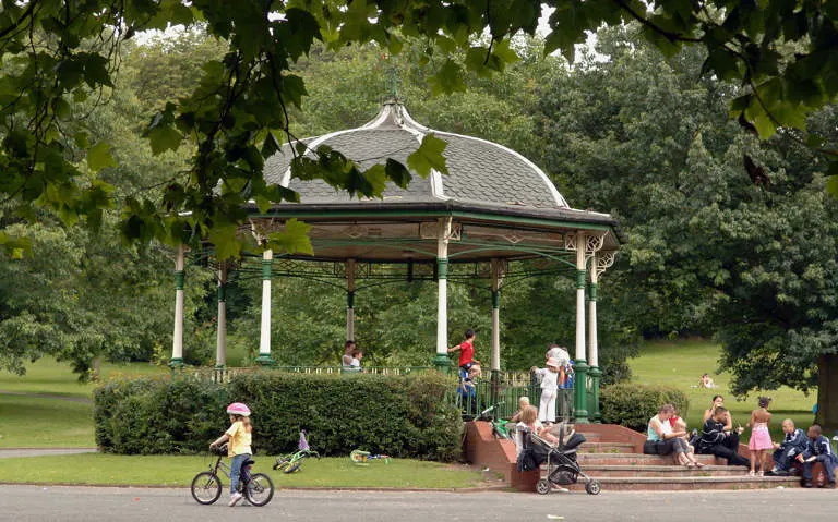 A Victorian bandstand with white columns and domed roof surrounded by trees, with families and children playing and relaxing in the park around it.