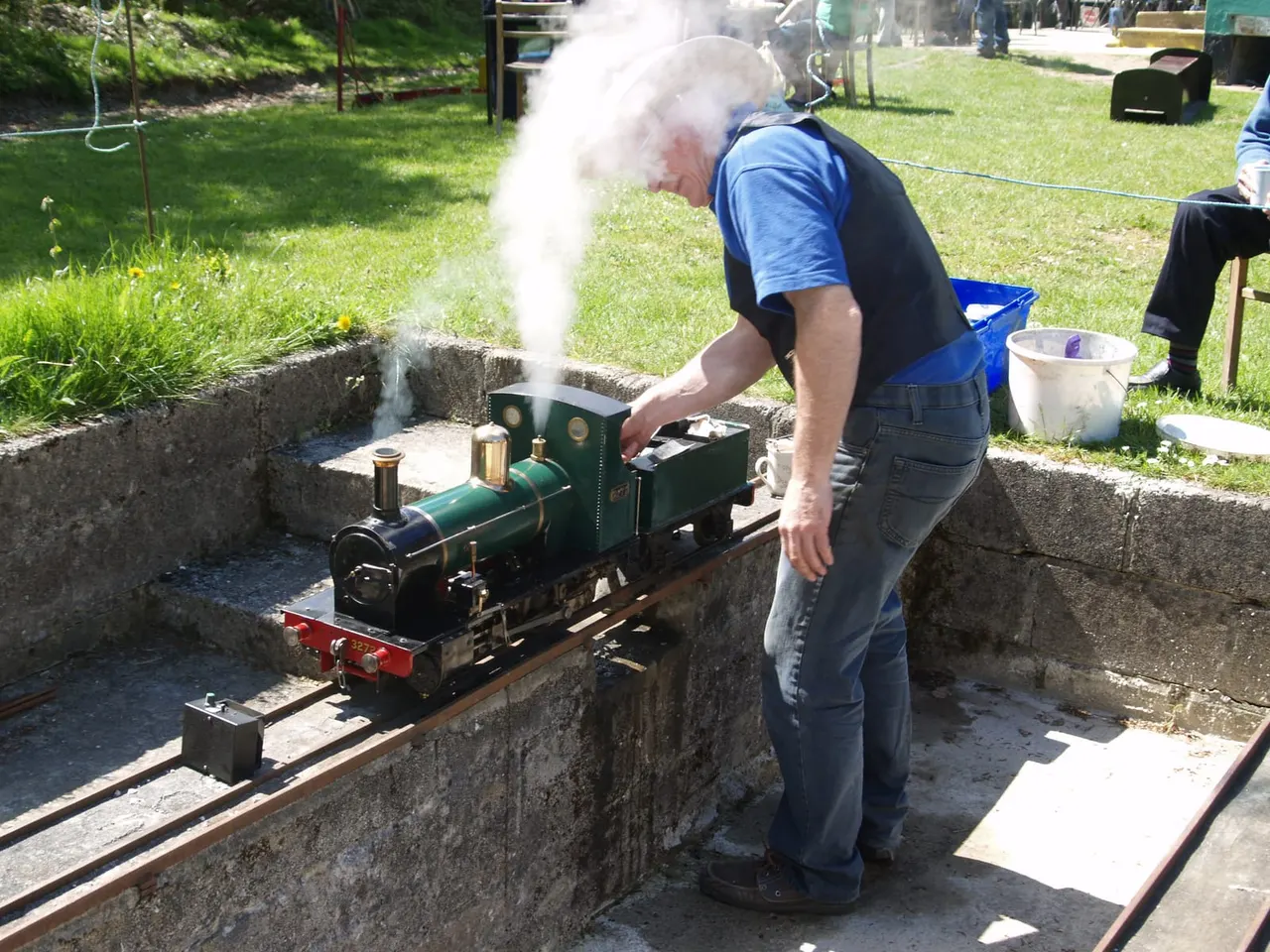 Man operating a miniature steam locomotive that is actively producing steam on an outdoor railway track.