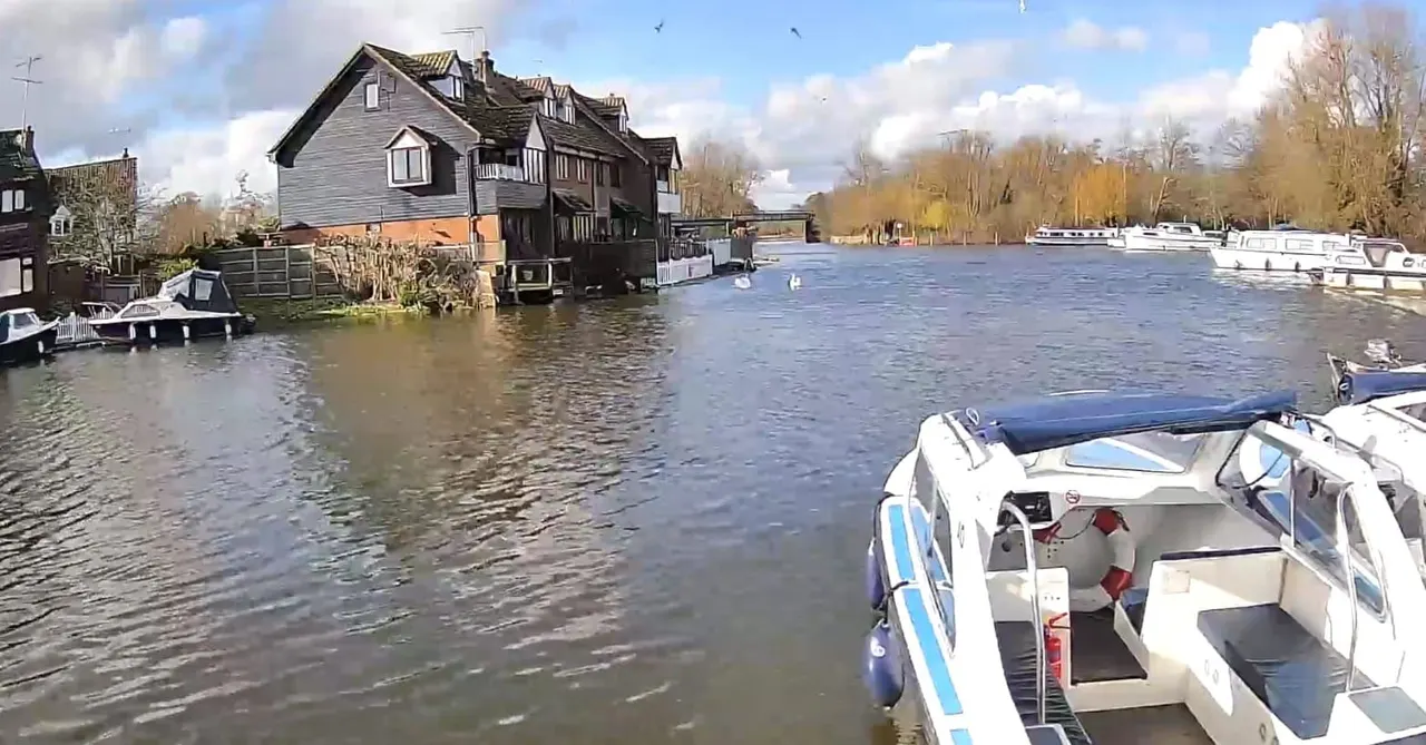 Moored motorboats on a river beside a historic waterfront house with bare winter trees and other vessels in the distance.