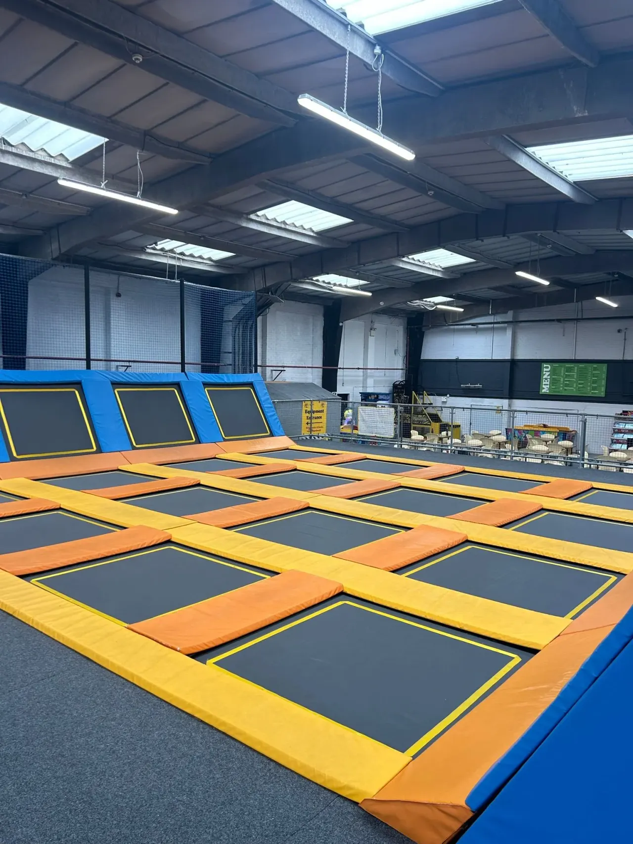 Indoor trampoline park with rows of interconnected black trampolines with orange and yellow padding.