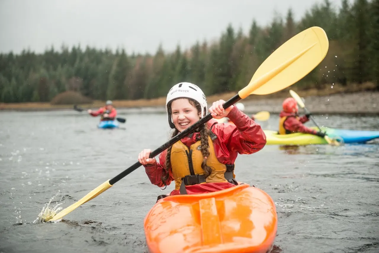 Child in safety helmet and lifejacket paddles an orange kayak on a lake with coniferous forest behind and other kayakers nearby.