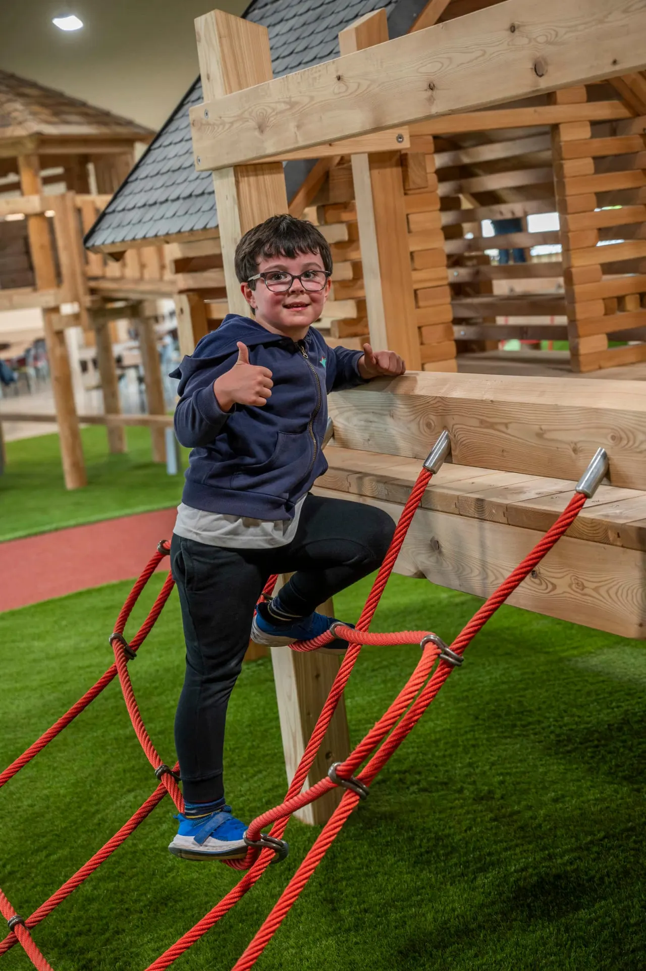 Child climbing on wooden play structure with red ropes in indoor play area.