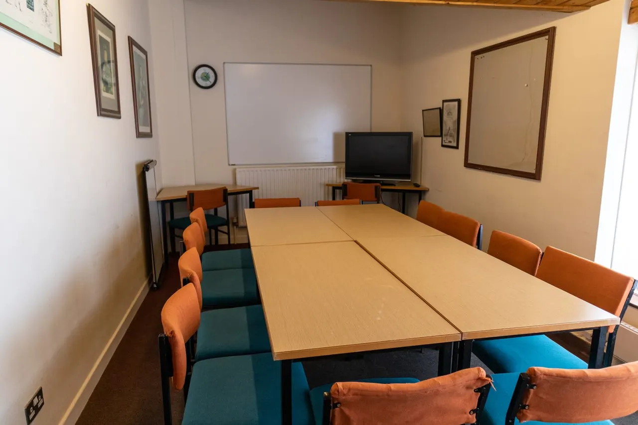 Empty meeting room with long wooden table, orange chairs, whiteboard, television and framed pictures on walls.