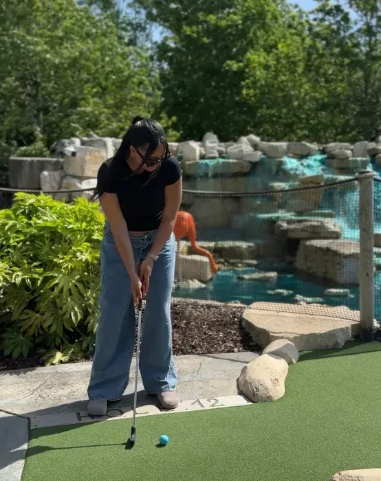 Woman playing mini golf on artificial green with crocodile enclosure visible behind fencing.