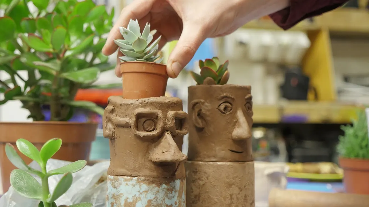 Person holding a small succulent in a terracotta pot above decorative face-shaped clay planters containing succulents.