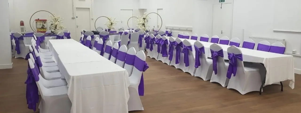 Event space decorated with long white-clothed tables and white chairs tied with purple ribbons, featuring circular floral arrangements.
