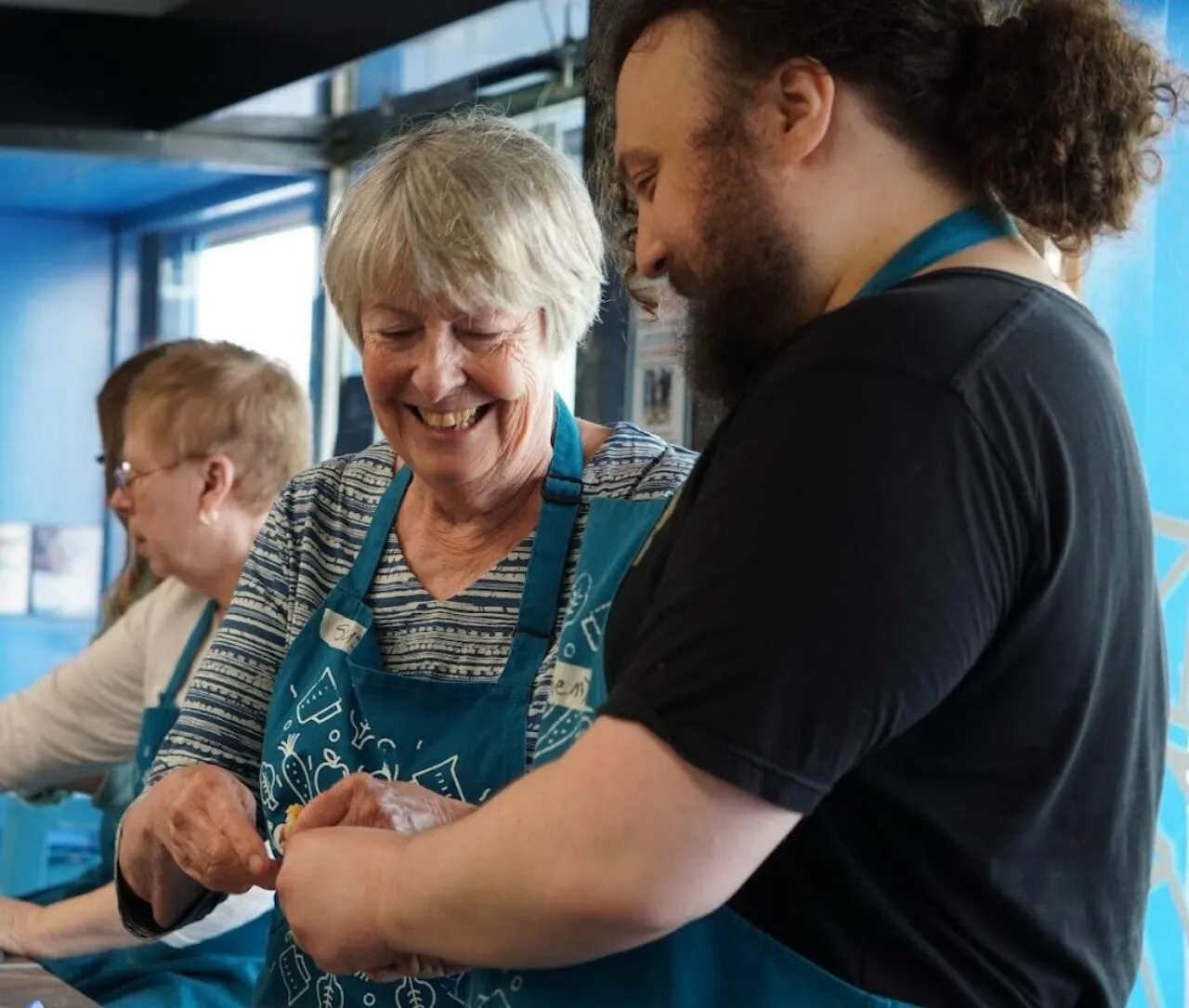 An older woman in a blue apron laughs whilst a bearded man guides her hands during a cooking activity, with others nearby.